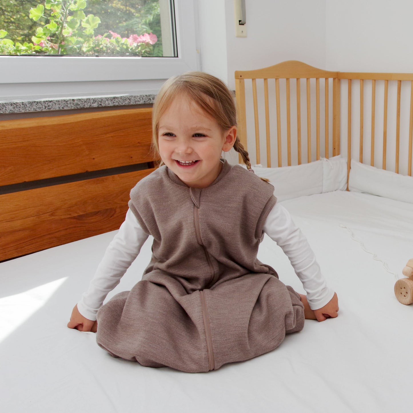A child wearing a brown wool sleep sack is sitting on a bed in a bedroom.