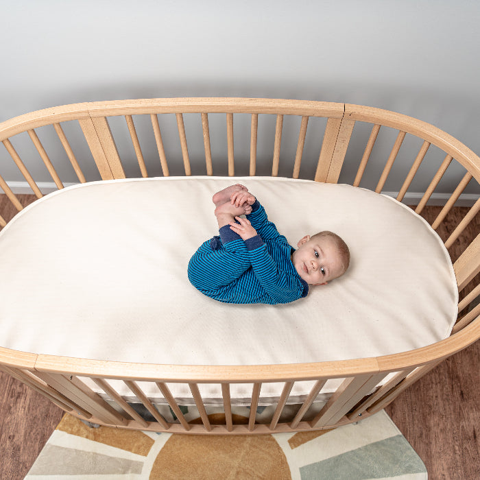 Baby lying in a wooden crib with a white mattress in a room.